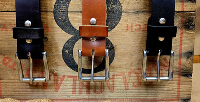 3, 1.5" plain leather belts in dark brown, tan and black leather with square silver buckles shown draped over an old brown wooden box with burlap textured fabric