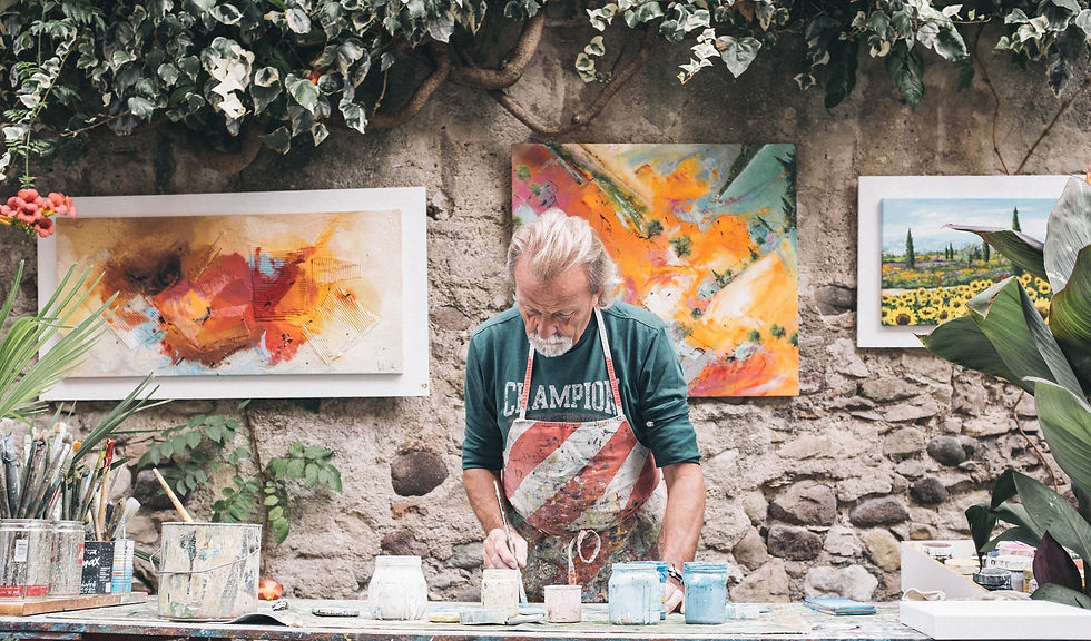 A Roman artisan painting at an outdoor workshop in Rome, Italy, with his paintings displayed behind him on an ancient stone wall covered in vines.
