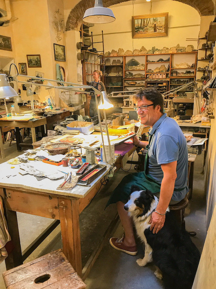 man working at a big desk with a dog beside him inside an artisan workshop for pietra dura artworks