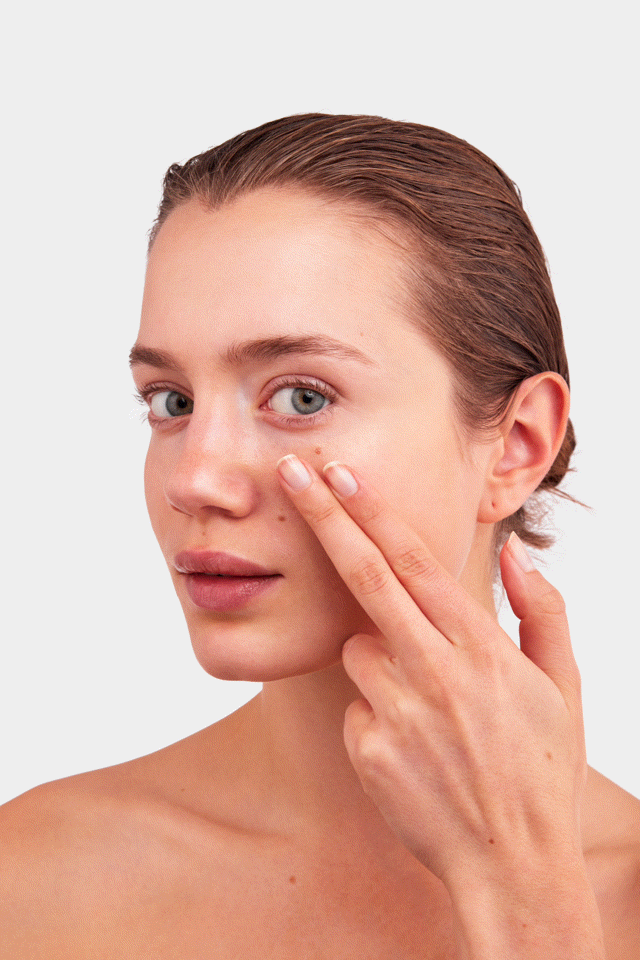 Close-up of a woman on a white background touching the skin under her eye, with a calm, focused expression.