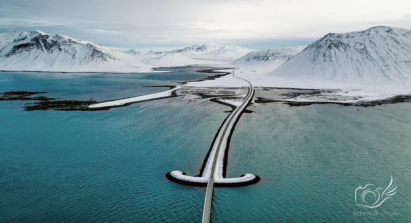 Landschaftsaufnahme einer Straße über türkisblaues Wasser zwischen schneebedeckten Bergen