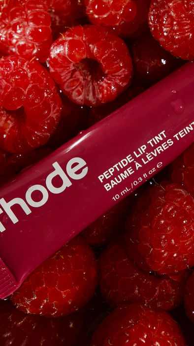 Rhode peptide lip tint placed among fresh raspberries, beauty product photography.