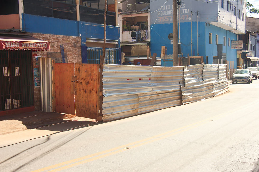 Na extremidade esquerda da foto, o mercadinho de Graciele, ao lado dele a papelaria e residência de Juliana. Foto: Douglas Júnio. #ParaTodosVerem: Foto de uma rua com um canteiro de obras cercado por tapumes metálicos e um portão de madeira. À esquerda, há um pequeno mercado; ao fundo, prédios comerciais e residenciais. Alguns carros estão estacionados na via, em um dia ensolarado.