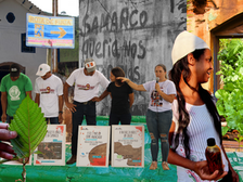 A imagem é uma montagem fotográfica que reúne diferentes cenas relacionadas à memória e resistência após o rompimento da barragem de Fundão, em Mariana (MG). Há pessoas com camisetas de protesto, cartazes denunciando crimes da Samarco, uma parede pichada com a frase: “A Samarco queria nos matar, mas Jesus nos salvou”, uma placa de “rota de fuga”, além de cenas de diálogo comunitário e cuidado com a natureza, representados por uma horta e uma folha verde em destaque.