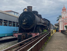 Imagem da Maria fumaça, trem famoso e histórico da cidade Mariana. O fundo é azul, e possui uma construção rosa ao lado.