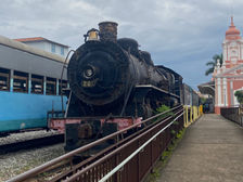 Imagem da Maria fumaça, trem famoso e histórico da cidade Mariana. O fundo é azul, e possui uma construção rosa ao lado. 