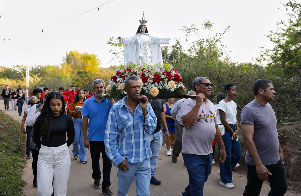 #ParaTodosVerem: Procissão religiosa ao ar livre durante o dia. Quatro homens carregam um andor com a imagem de uma santa vestida de branco e coroada. Na parte de cima do andor há um grande arranjo de flores nas cores vermelha, salmão e branca, que envolvem a parte de baixo da imagem. Ao redor, a comunidade do subdistrito atingido caminha, em procissão, por uma estrada de terra, em meio à vegetação.