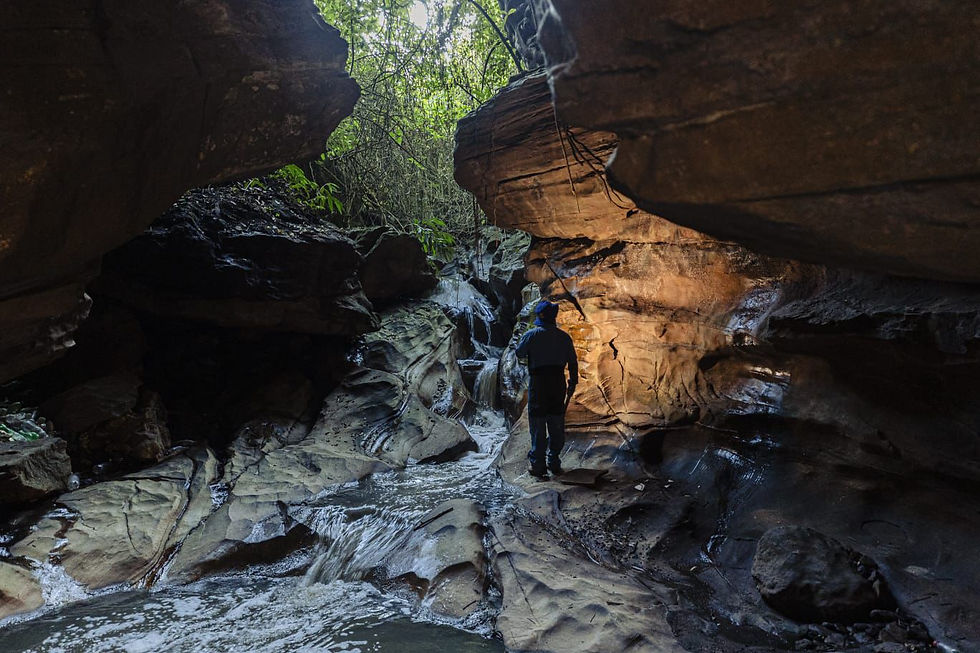 #ParaTodosVerem: Um homem de capacete e roupas escuras em uma pequena caverna esculpidas pela água. Um riacho corre sob seus pés e a luz do sol penetra entre as árvores no topo das rochas, iluminando parcialmente o interior úmido e sombreado da formação natural.