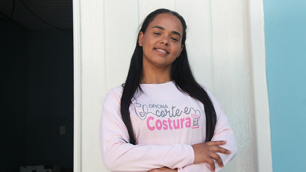 A imagem mostra uma mulher negra, sorrindo em frente à fachada da Oficina de Corte & Costura. Ela veste camiseta rosa com o nome do projeto estampando.