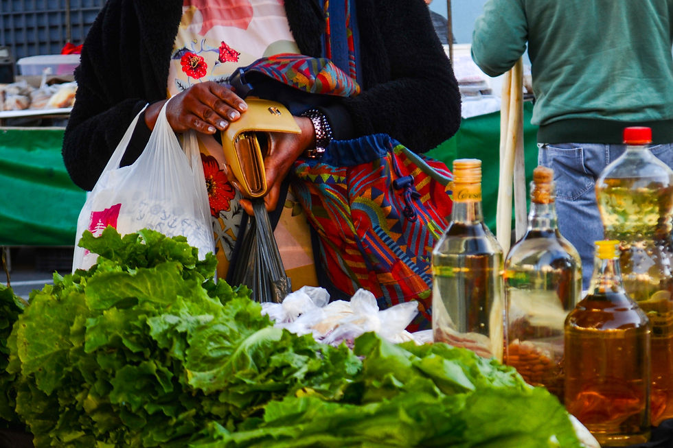 #ParaTodosVerem: A foto foca nas mãos de uma mulher em uma feira, mostrando o momento em que ela paga suas compras. No primeiro plano, há um cacho de folhas verdes e várias garrafas com líquido dourado. A mulher, que veste uma blusa florida e um casaco preto, segura uma carteira aberta, enquanto carrega uma sacola plástica e uma de tecido.