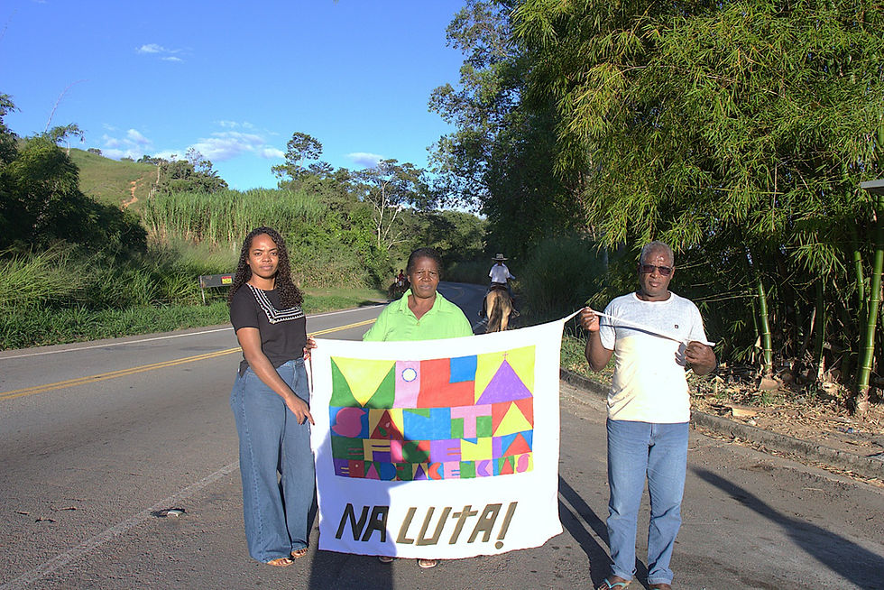 Daiane (esquerda), Neusa e João Carlos com a bandeira da Associação Vila Santa Efigênia e Adjacências na MG 262. Foto: Tainá Quirino. #ParaTodosVerem: No lado esquerdo está a Daiane,  mulher negra de cabelo cacheado longo e solto, veste calça jeans e blusa preta (à esquerda), ao centro está Neusa mulher idosa negra, usando blusa verde neon, e, à direita, está o João Carlos homem negro adulto estindo blusa branca e calça jeans.. Os três seguram a bandeira da Associação, que contém símbolos coloridos que formam o nome da comunidade Vila Santa Efigênia e Adjacências e embaixo a frase “ NA LUTA!”, às margens da rodovia MG 262. 
