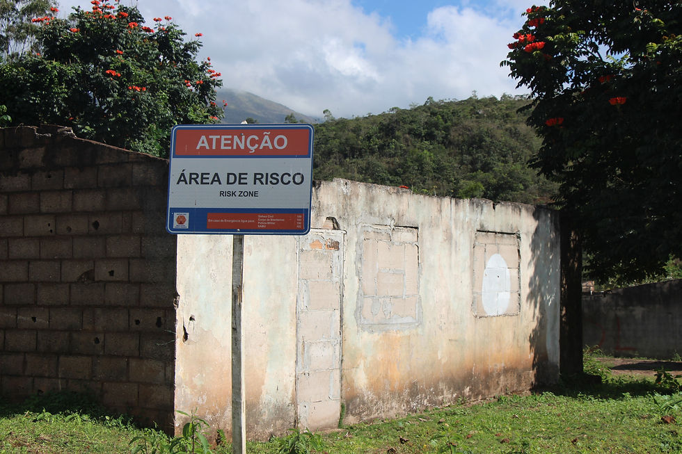 Antiga moradia tem suas portas e janelas fechadas por paredes para evitar invasões e vandalismo. Foto: Ana Giulia Bento. #ParaTodosVerem: Placa “Atenção: Área de Risco” diante de ruínas de construção sem telhado. Ao fundo, vegetação, árvores floridas e uma serra coberta por nuvens.