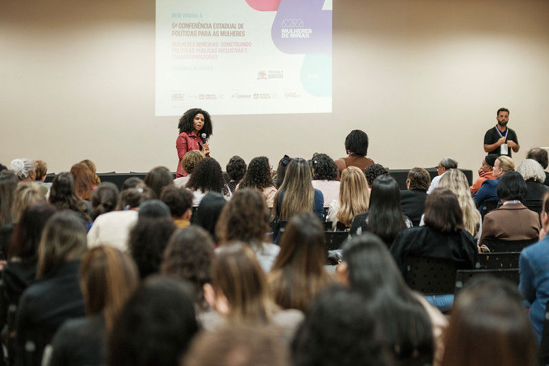 #PraTodosVerem: Na imagem, uma mulher fala ao microfone para um auditório cheio de mulheres e alguns homens, de costas para a câmera. No telão ao fundo, está o painel da 5ª Conferência Estadual de Políticas para as Mulheres e o projeto Mulheres de Minas.