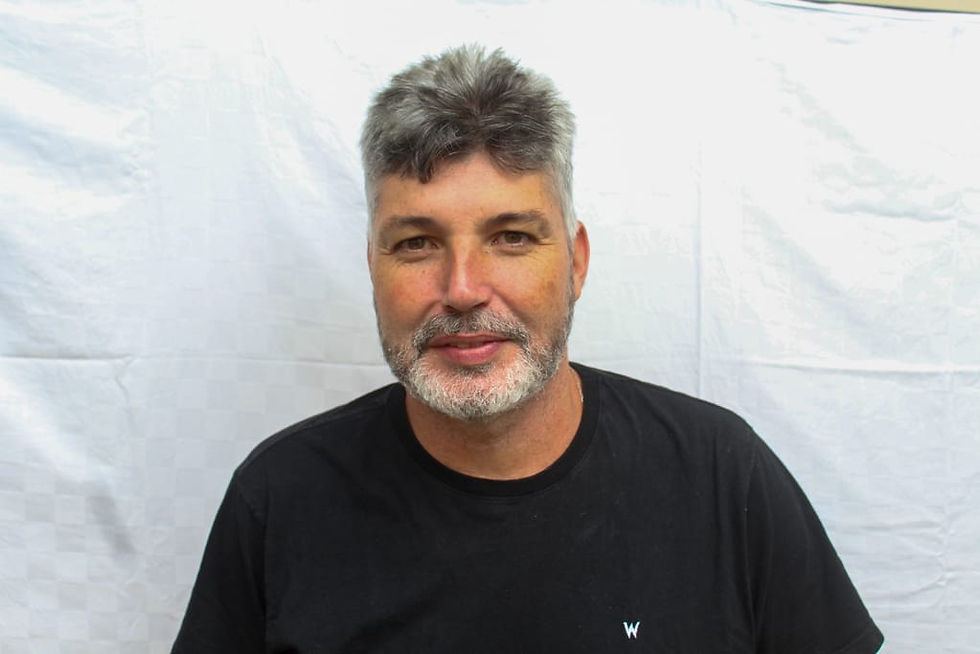 #ParaTodosVerem: Homem de cabelos grisalhos, barba branca, pele clara, vestido com uma camisa preta lisa e com uma expressão leve e um pequeno sorriso. Ele está sentado, em primeiro plano, com um fundo branco atrás.