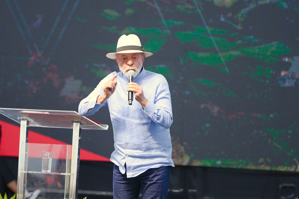 O presidente Lula, um homem de chapéu claro e camisaEm seu discurso, o presidente critica o acordo inicial, costurado pelas mineradoras nos governos Temer e Bolsonaro. azul fala ao microfone em um púlpito de acrílico. Ao fundo, um telão mostra uma paisagem montanhosa