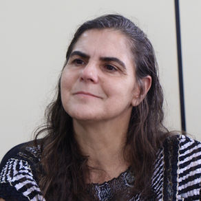 #ParaTodosVerem: A imagem mostra uma mulher com mais de 50 anos, branca, de cabelo preto e longo, partido para a lateral esquerda. Ela usa uma blusa preta com detalhes brancos e está sorrindo.