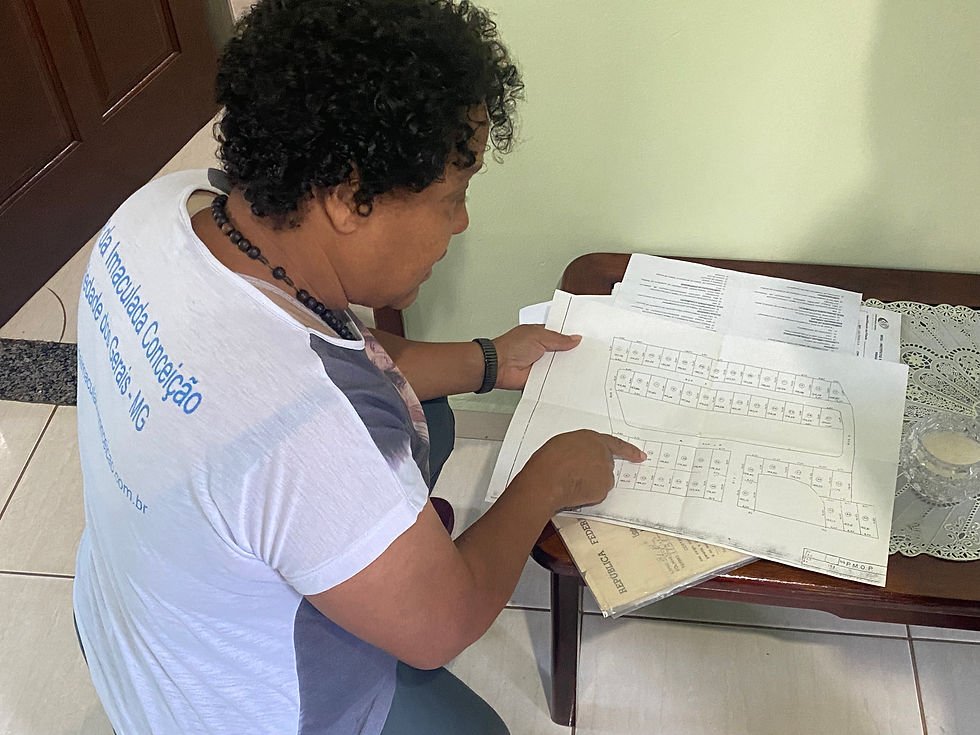 #ParaTodosVerem: Mulher de cabelo cacheado, usando uma blusa branca com estampa e um colar, ajoelhada no chão, olhando um plano urbanístico. Ela está apontando para o documento, mostrando detalhes de um projeto.
