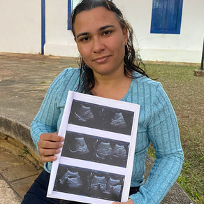 Uma mulher sentada ao ar livre, em frente a uma parede branca com detalhes em azul. Ela veste uma blusa azul clara e calça jeans, e segura nas mãos um papel com imagens de ultrassom em preto e branco, mostrando quatro registros médicos. Ela olha diretamente para a câmera com expressão serena.