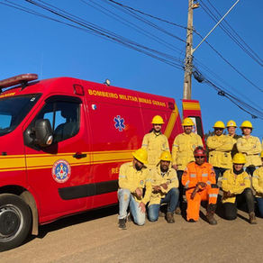 Em uma estrada sob céu azul e limpo, um grupo de pessoas posa ao lado de uma viatura vermelha do Corpo de Bombeiros Militar de Minas Gerais. Todos os brigadistas vestem jaquetas e capacetes amarelos, enquanto o bombeiro militar ao centro está com uniforme laranja. Todos estão sorrindo, parte está agachada e parte está em pé. Ao fundo, há postes de energia e fiação elétrica.