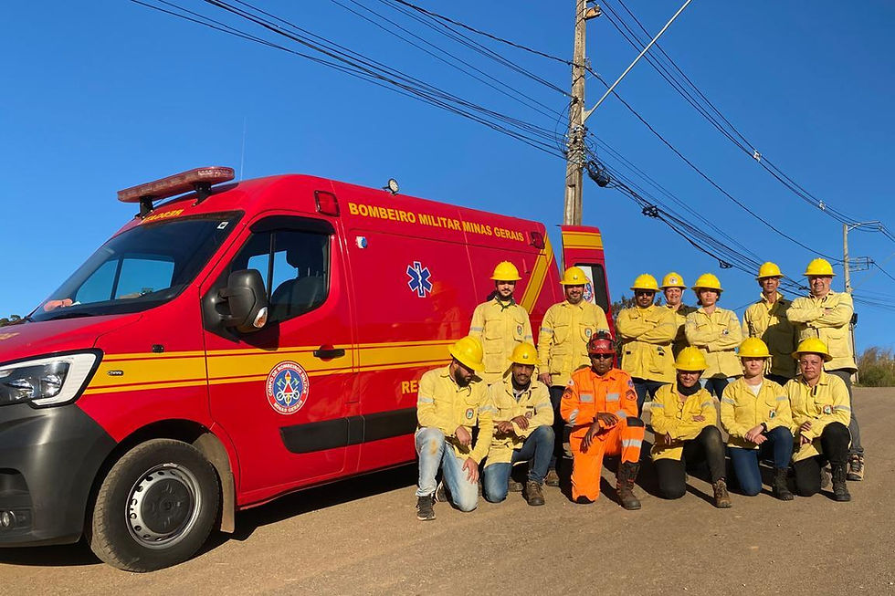 Em uma estrada sob céu azul e limpo, um grupo de pessoas posa ao lado de uma viatura vermelha do Corpo de Bombeiros Militar de Minas Gerais. Todos os brigadistas vestem jaquetas e capacetes amarelos, enquanto o bombeiro militar ao centro está com uniforme laranja. Todos estão sorrindo, parte está agachada e parte está em pé. Ao fundo, há postes de energia e fiação elétrica.