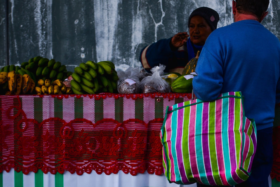 #ParaTodosVerem: A foto mostra um cliente de costas, carregando uma sacola de compras grande e listrada, em frente a uma barraca de frutas. A sacola é vibrante, com listras verticais em tons de rosa, verde, roxo e azul. O cliente, que veste uma blusa azul escura, interage com a vendedora, que tem um gorro e está atrás de uma banca com toalha de renda vermelha e babados listrados em verde e branco. Sobre a banca, há cachos de bananas verdes e maduras, além de sacos com outros produtos.