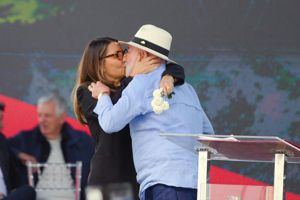 O presidente Lula, usando chapéu e uma mulher, a primeira-dama Janja, se beijam em um palco, com uma grande tela ao fundo exibindo uma paisagem.