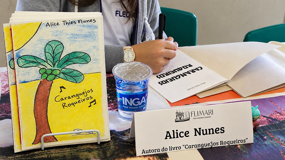 #ParaTodosVerem: Sobre uma mesa, há uma cópia do livro “Caranguejos Roqueiros”, de Alice Nunes, exposto em um suporte. A capa traz o desenho de um coqueiro na praia. Ao lado, há um copo de água mineral e uma placa de identificação da autora mirim. Ao fundo, Alice está segurando uma caneta preta e autografando seu livro.