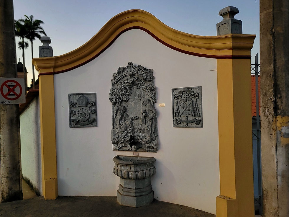 Para todos verem: Um chafariz histórico de parede, com estrutura em pedra esculpida e moldura pintada de amarelo e branco. No centro, há um grande painel em pedra com relevos, ladeado por dois painéis menores, também esculpidos. Na base central, está a bica d’água sobre uma pequena pia de pedra.