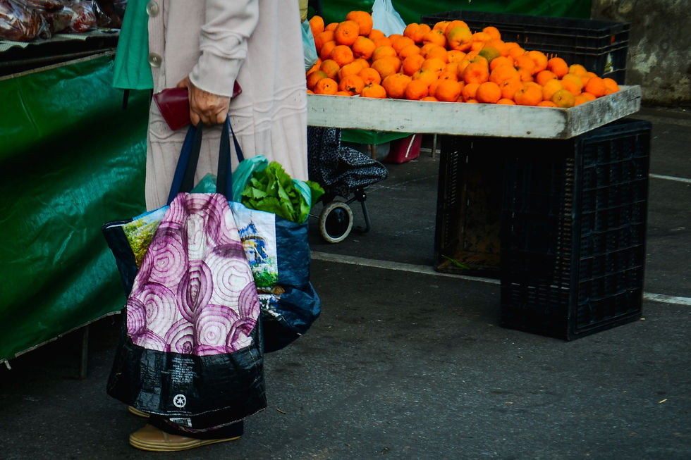 #ParaTodosVerem: A foto mostra o tronco e as pernas de uma pessoa em uma feira, carregando várias sacolas. A pessoa, que está de lado, usa uma blusa ou casaco longo de cor creme ou bege. Nas mãos, ela carrega diversas sacolas, sendo uma delas um saco de pano com uma estampa roxa e branca que lembra anéis de cebola. A pessoa usa sapatos de cor clara e também carrega uma pequena bolsa vermelha. Ao lado, há uma banca de feira com uma grande quantidade de laranjas em uma caixa de madeira. O chão é de asfalto.