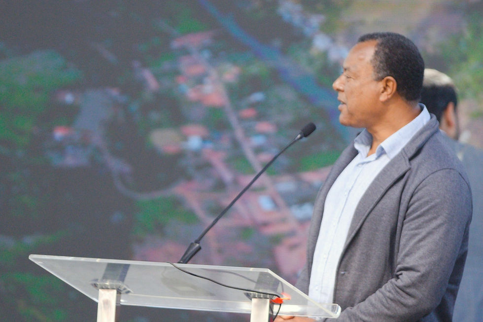 Mauro da Silva, um homem negro vestido com uma camisa social de cor clara e um blazer cinza, fala em um púlpito de acrílico com microfone, em frente a um telão que exibe uma paisagem.