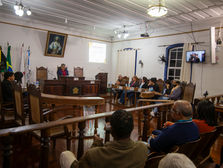 A imagem mostra uma audiência pública acontecendo em um salão da Câmara Municipal. As pessoas presentes estão sentadas em cadeiras, organizadas em fileiras. Na parte da frente, há uma mesa grande com várias pessoas que estão representando instituições e serviços que diariamente trabalham com pessoas em situação de rua.O local tem paredes claras, teto branco e chão de madeira escura. Há bandeiras ao fundo e dois quadros pendurados na parede.