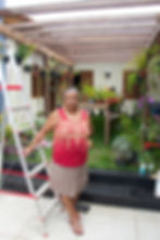 #ParaTodosVerem: Senhora, de cabelos grisalhos presos, usando blusa vermelha com estampa amarela, saia cinza e chinelo. Ela está em pé e apoiada em uma escada de alumínio. Ao fundo, janelas da parte interior da casa e plantas.