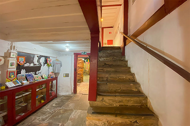 Foto do subsolo do Museu Casa Guignard. A esquerda está uma escada com corrimão que leva ao térreo e à direita tem um balcão de vidro de uma lojinha de lembranças como quadros, bandeiras de santos e camisas. 