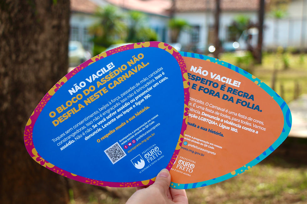 #ParaTodosVerem: A imagem mostra uma mão segurando dois leques de papel nas cores azul escuro e laranja. Os leques possuem frases de aviso sobre o assédio sexual e a LGBTQIAPN+ durante o período de Carnaval. No leque azul, mais à frente, lê-se: “Não Vacile! O bloco do assédio não desfila neste carnaval”. Abaixo há um texto sobre o que é assédio, incentivando a denúncia, o leque conta também com um qr code com a programação do carnaval e a logo da Prefeitura de Ouro Preto.