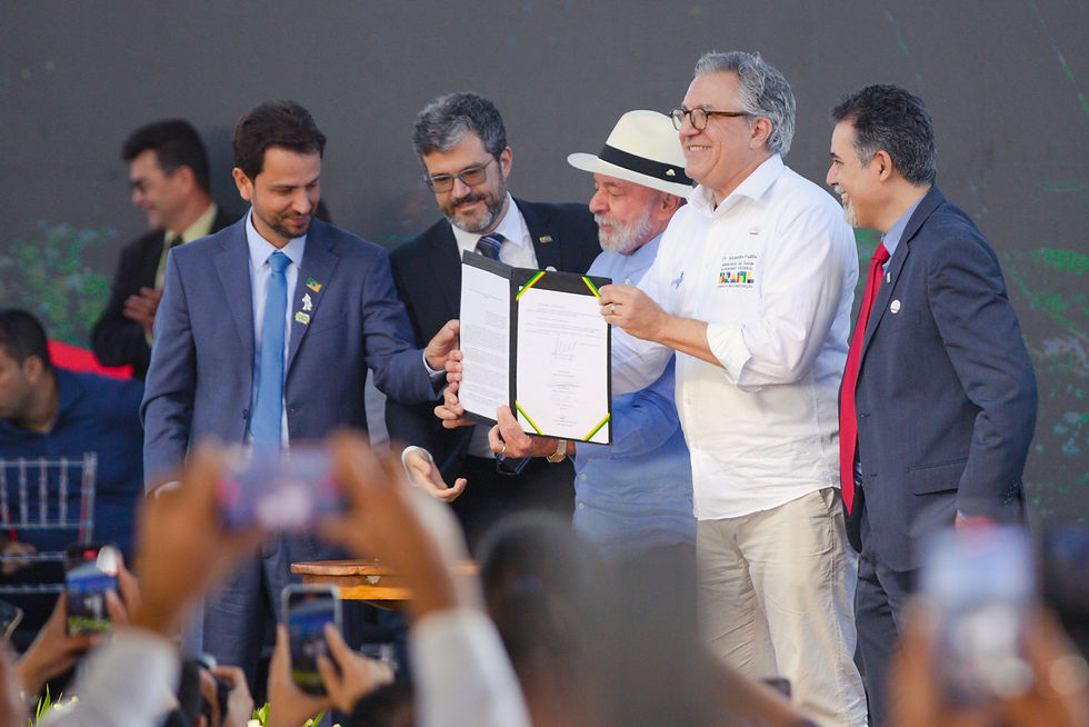 A imagem mostra cinco homens no palco de um evento, todos sorrindo. O presidente Lula, ao centro, com chapéu branco e camisa clara, segura um documento aberto. Os outros quatro, em trajes sociais, estão ao redor dele, observando o documento. No fundo, um telão exibe uma paisagem.