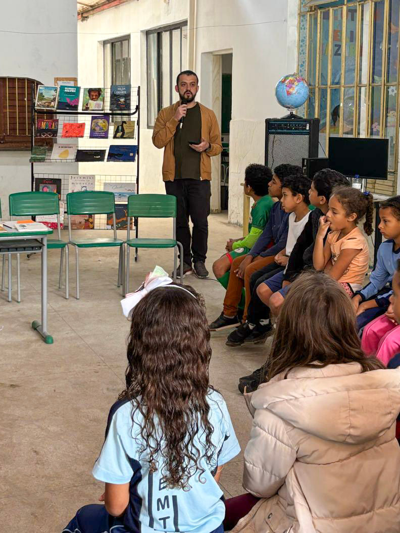 #ParaTodosVerem: Um homem de barba, com casaco marrom-claro, fala ao microfone para um grupo de crianças e adolescentes sentados no chão, em uma área coberta. Ele está do lado de uma estante com livros coloridos. As crianças, de diversas etnias, olham atentamente. A cena sugere uma atividade de contação de histórias ou leitura em um ambiente escolar.
