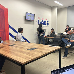 Uma sala com paredes brancas com pessoas e sentadas em cadeiras ao redor de uma mesa retangular de madeira, participando de uma apresentação. Há uma mulher ao fundo com camisa listrada preta e branca que fala para o grupo enquanto a tela na parede exibe slides. Algumas pessoas estão com notebooks no colo ou sobre a mesa.