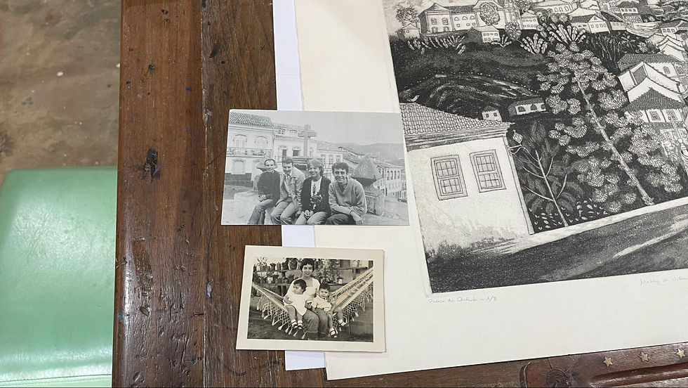 #ParaTodosVerem: Foto feita de cima mostra uma mesa de madeira com duas fotografias antigas em preto e branco e uma gravura. Em uma das fotos, quatro jovens estão sentados em um muro, com construções históricas ao fundo. Na outra, uma mulher segura duas crianças sentadas em uma rede. Ao lado, há uma gravura em preto e branco de um casario colonial em meio a uma paisagem montanhosa. À esquerda, aparece parte de uma cadeira verde.