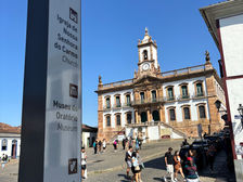 Fotografia da Praça Tiradentes, em Ouro Preto, com destaque para um totem de sinalização em primeiro plano, que traz indicações bilíngues em português e inglês para pontos turísticos próximos. Ao fundo, vê-se o Museu da Inconfidência, com sua fachada colonial e torre do relógio, além de turistas caminhando pelo calçamento de pedra.