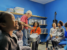 Descrição: #ParaTodosVerem: duas crianças sorriem dentro de uma sala com instrumentos musicais de percussão e caixas organizadoras de outros materiais da aula. Ao fundo, aparece um senhor e outras crianças.