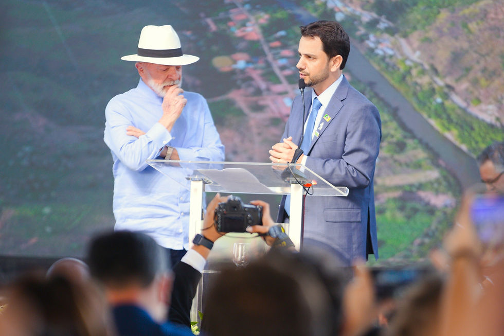 A imagem mostra dois homens em um palco: à esquerda,o presidente Lula, usando chapéu e camisa azul clara, e à direita, o prefeito Juliano Duarte, de terno azul, falando em um púlpito com microfone. Atrás deles, um telão exibe paisagem com rio e montanhas.
