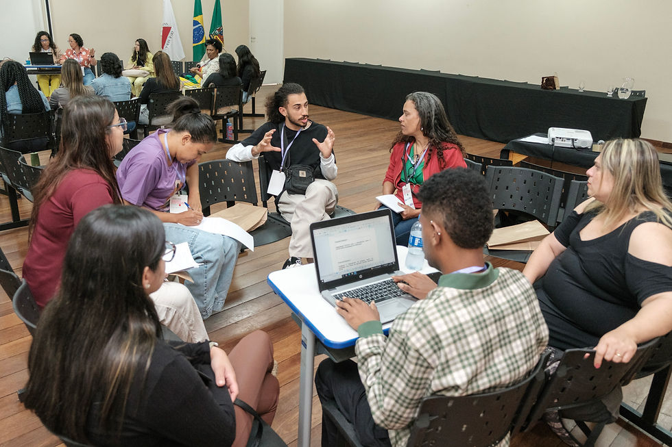 #PraTodosVerem:Fotografia em ambiente interno mostra um grupo de sete pessoas sentadas em círculo, participando de uma roda de conversa durante a conferência. No centro, um homem de camisa preta gesticula enquanto fala, sendo escutado atentamente por uma mulher de blusa vermelha à sua frente. Outras pessoas fazem anotações. Ao fundo, há mais participantes em roda. Atrás, estão as bandeiras do Brasil, de Minas Gerais e de Mariana.