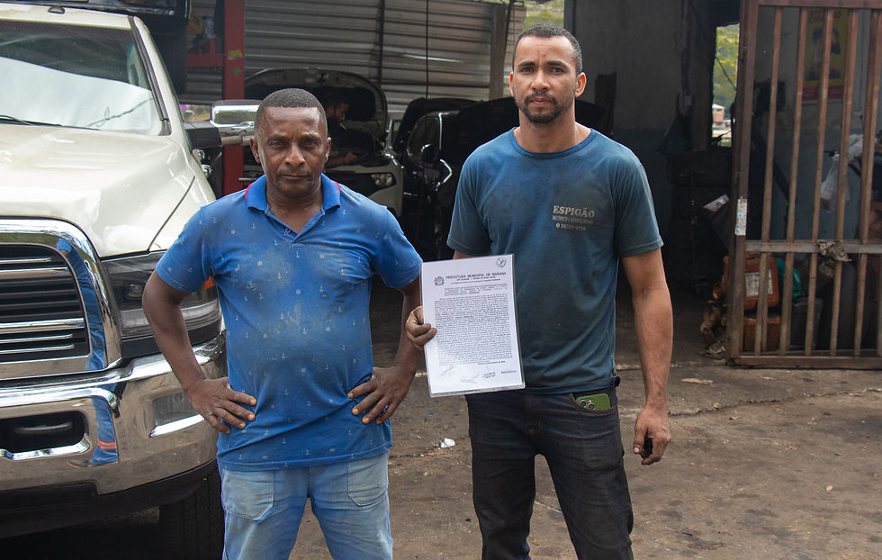 Marcelo França (à esquerda) e João Hernandes mostram escritura particular de doação (termo de posse) de imóvel, assinado pelo ex-prefeito Celso Cotta. Foto: Fernanda França. #ParaTodosVerem - Dois homens exibem documento de posse em frente a uma mecânica. Na camisa de um deles, está escrita a palavra “Espigão”.