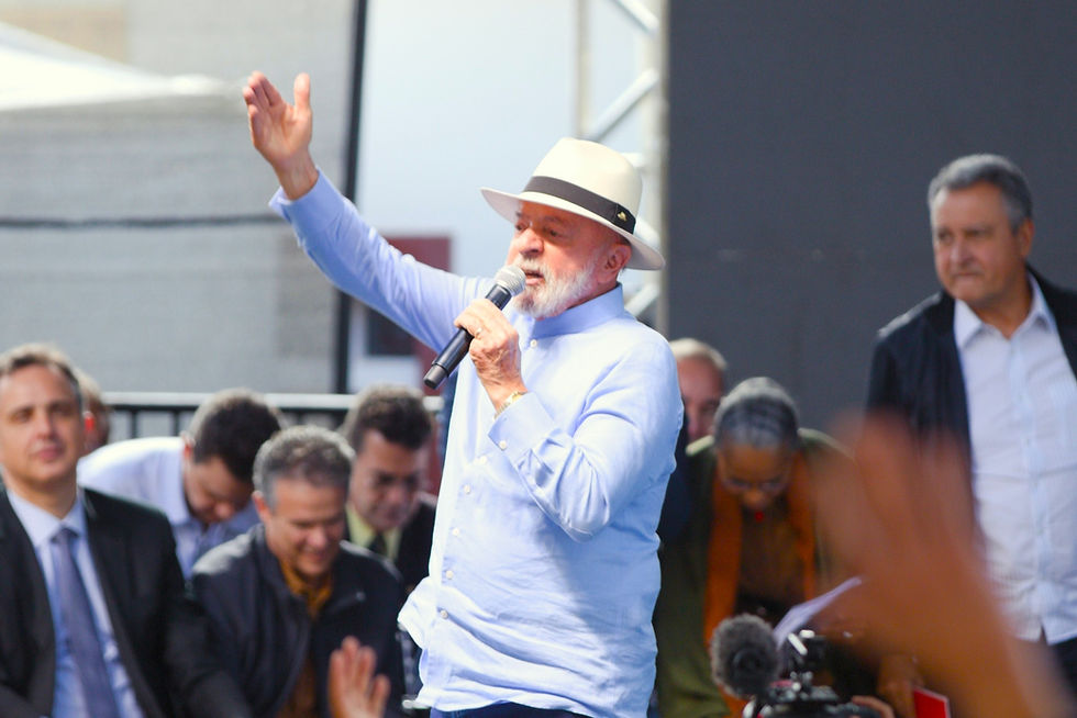 Cena de um evento público, com o presidente Lula, um homem de pele clara no centro, vestindo uma camisa social azul clara de manga comprida, calça jeans escura e um chapéu branco. Ele está em um palco, segurando um microfone com a mão esquerda e estendendo o braço direito para cima, com a palma da mão aberta, como se estivesse cumprimentando ou interagindo com a plateia.