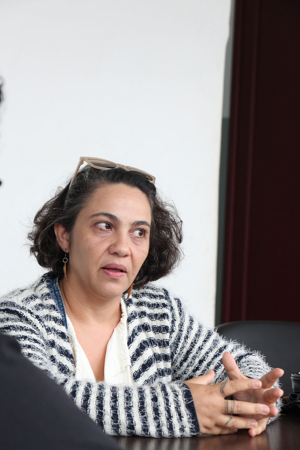 #ParaTodosVerem: Na imagem, uma mulher de cabelos pretos, a diretora Luciane Trevisan, veste um casaco branco com listras pretas. Possui os óculos posicionados no topo da cabeça e fora dos olhos. Ela olha para o lado e gesticula com as mãos sobre a mesa. O fundo da foto é uma parede branca.