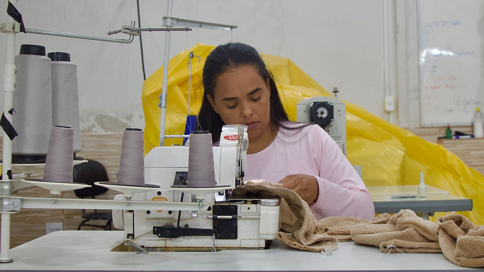 Mulher negra, aparece sentada à máquina de costura, concentrada na confecção de um casaco de pelúcia branco. Ela usa camiseta rosa e está dentro da Oficina de Corte & Costura, com tecidos e materiais da cor marrom ao redor.