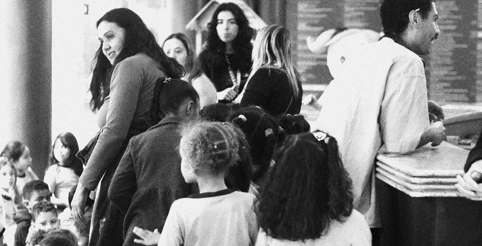 #PraTodosVerem: Foto em P&B de várias crianças em pé organizadas em fila em um saguão, acompanhadas por adultos.