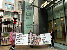 #Para todos verem: Três mulheres estão em pé, lado a lado, segurando uma grande faixa em frente ao prédio da Rolls Building – Royal Courts of Justice, em Londres. A faixa mostra fotos de 19 pessoas e os dizeres, em inglês: "These are the victims of the Mariana Dam disaster. Will they get justice?" (“Essas são as vítimas do desastre da barragem de Mariana. Elas terão justiça?”).