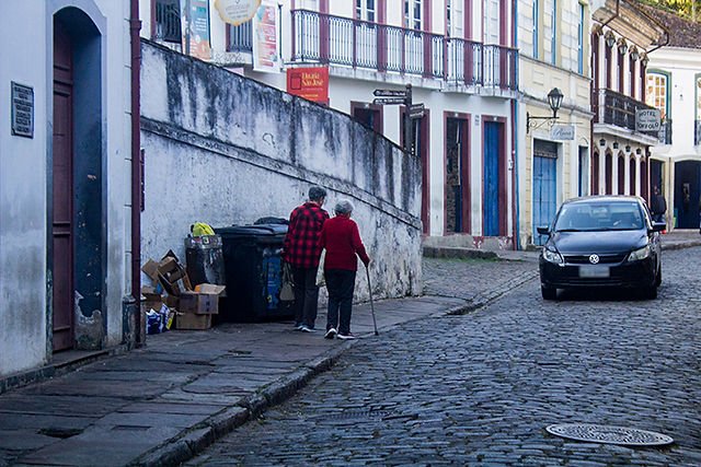 Foto de uma rua de pedra. Na calçada, é possível ver duas mulheres com cabelo curto e grisalho, que aparentemente estão de braços dados, uma delas segura uma bengala. Ao fundo estão casas características do centro histórico de Ouro Preto. Todas brancas com detalhes em azul ou vermelho escuro, de dois andares e com sacadas. Passa um carro na rua. 