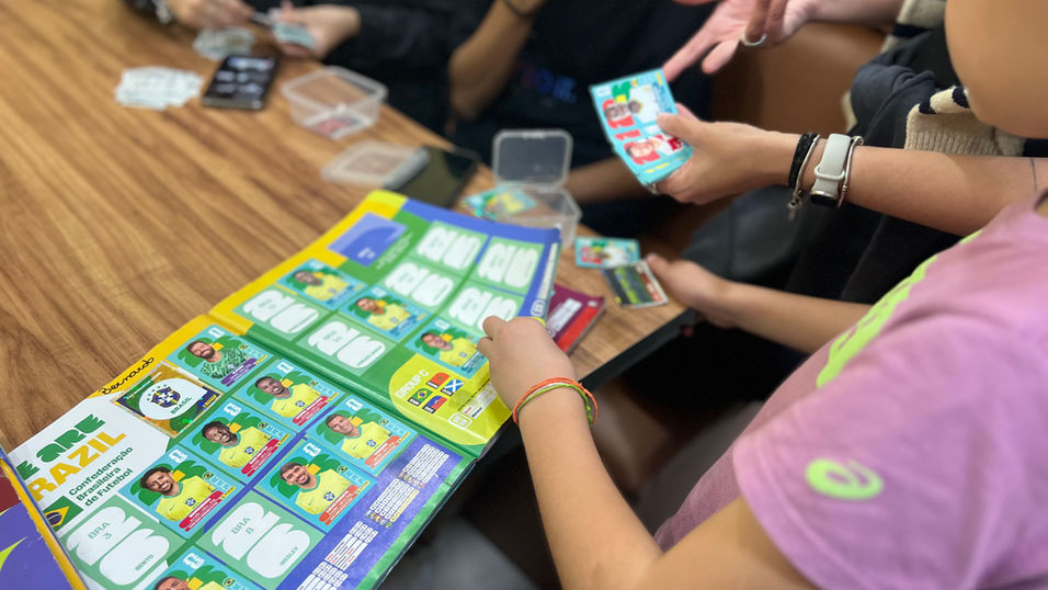 Grupo de pessoas participa de uma troca de figurinhas da Copa do Mundo 2026 ao redor de uma mesa. Uma criança, enquadrada de lado e com os rosto desfocado segura o álbum aberto, enquanto os demais organizam e trocam cromos em um momento de convivência. O foco e destaque da foto estão no álbum e nas mãos de algumas crianças sentadas ao redor de uma mesa de madeira marrom claro.